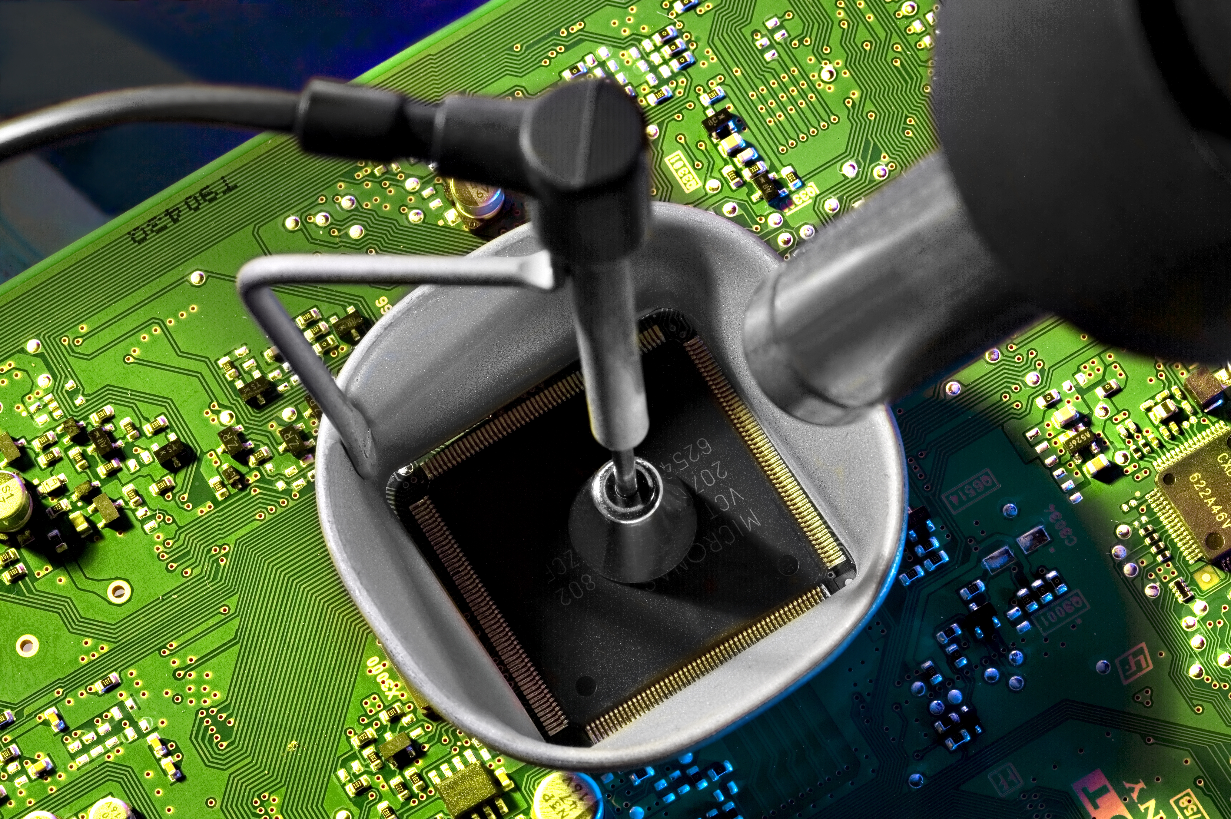 Photograph of an integrated circuit being desoldered from a green PCB using a hot-air rework station. The cylindrical hot-air nozzle is positioned directly above the chip and is delivering a focused stream of heated air onto the package to liquefy the solder underneath. The PCB is held in a clamp on a workbench. Other components on the surrounding board are visible. Forensic hot-air chip-off uses the same principle and equipment, paired with a board preheater for thermal control and vacuum tweezers to lift the chip the moment the solder reflows.